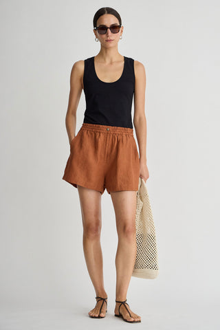 Woman wearing a black tank top and brown shorts, holding a beige bag on a light gray background