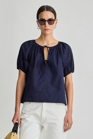 Woman wearing a navy blue blouse with puff sleeves and white pants on a light gray background