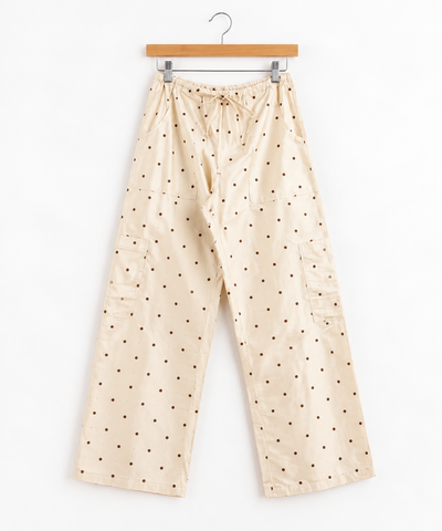 Cream-colored pants with a black polka dot pattern, featuring a drawstring waistband and side cargo pockets, hanging from a hanger, shown against a plain white background.
