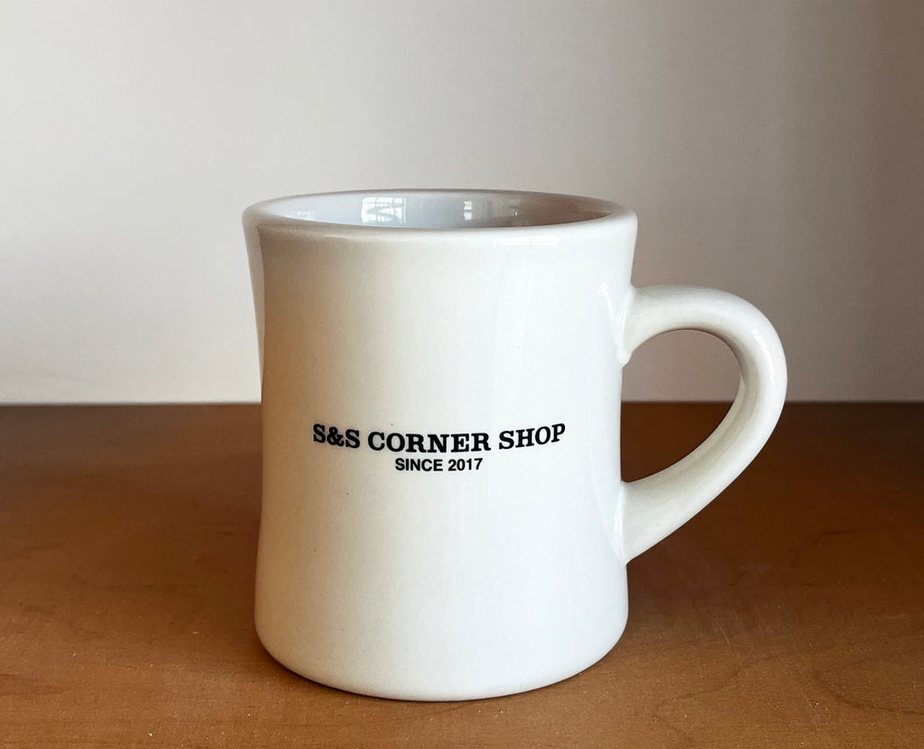 White mug with 'S&S Corner Shop Since 2017' text on a wooden surface.
