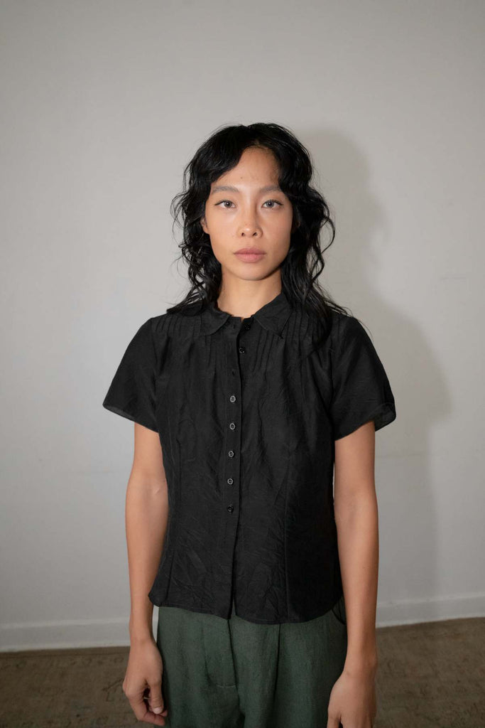 Woman wearing a Zii Ropa black short-sleeve button-down shirt with structured pintuck pleats along the shoulders, paired with dark green trousers.