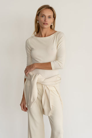 Model wearing a Boat Neck Top in Creme, paired with matching off-white pants and a sweater tied around the waist.