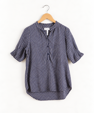 Apiece Apart navy blue and white gingham plaid short-sleeve shirt with a ruffled collar, a partial button placket, and ruffled sleeve cuffs, hanging on a wooden hanger.