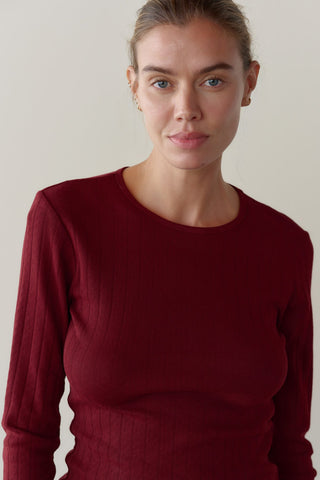 A close-up shot of a woman with her hair pulled back, wearing a fitted, burgundy-colored ribbed crewneck long-sleeve top against a neutral background.