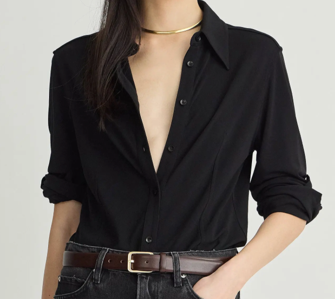 A woman wearing a black long-sleeve button-up shirt with rolled-up sleeves, paired with a brown belt and dark jeans, against a plain light background.