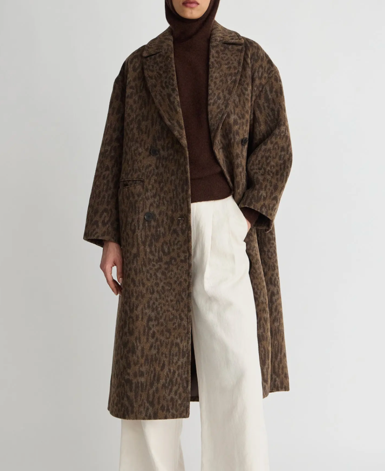 A model wearing a brown and black leopard print double-breasted coat over a dark brown turtleneck and white wide-leg trousers, shot against a plain light gray background.