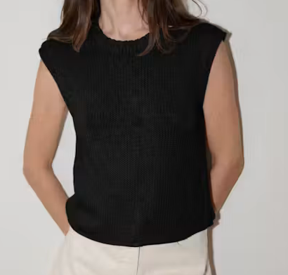 A woman wearing a sleeveless black ribbed knit top with a crew neckline, paired with off-white pants, shown against a plain light background.