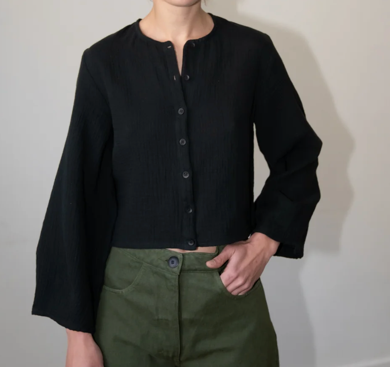 Woman modeling a black cropped long-sleeve button-up top with wide sleeves and a collarless round neckline, paired with olive green pants.