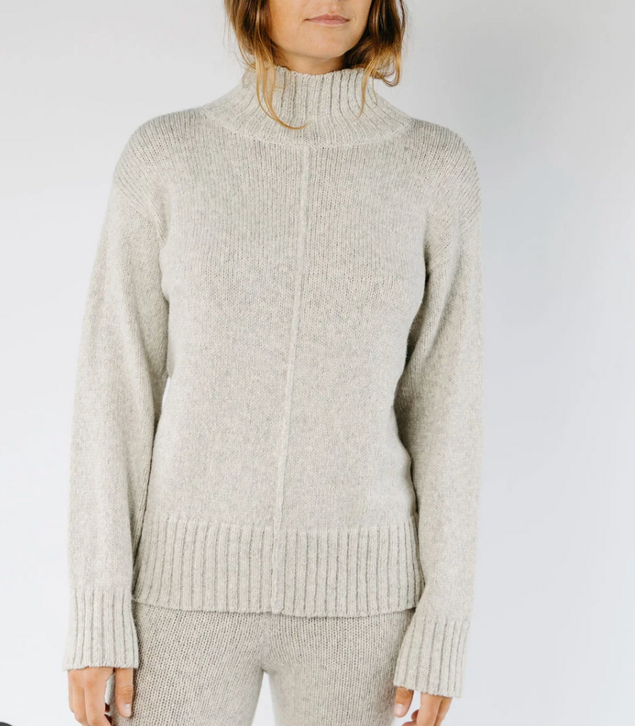 A woman wearing a light oatmeal-colored ribbed knit mock neck sweater and matching knit pants against a plain light gray studio background.