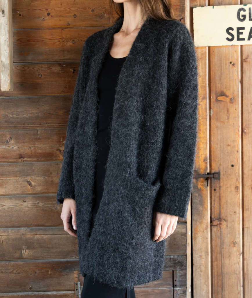 A woman wearing a dark charcoal gray fuzzy open-front cardigan over a black top, standing in front of a rustic wooden plank wall.