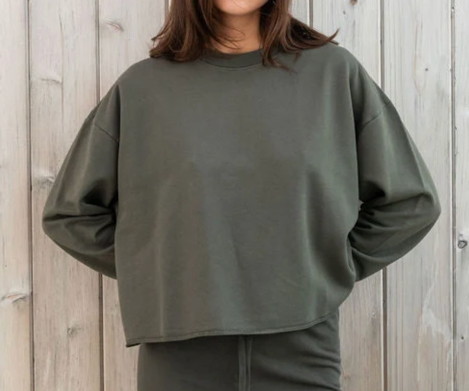 A person wearing an oversized crewneck sweatshirt in olive green, standing against a light gray wooden plank wall.