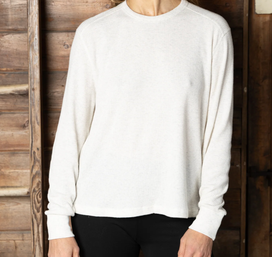 A person wearing a plain off-white long-sleeve crewneck sweater, paired with black pants, standing in front of a rustic wooden plank wall.