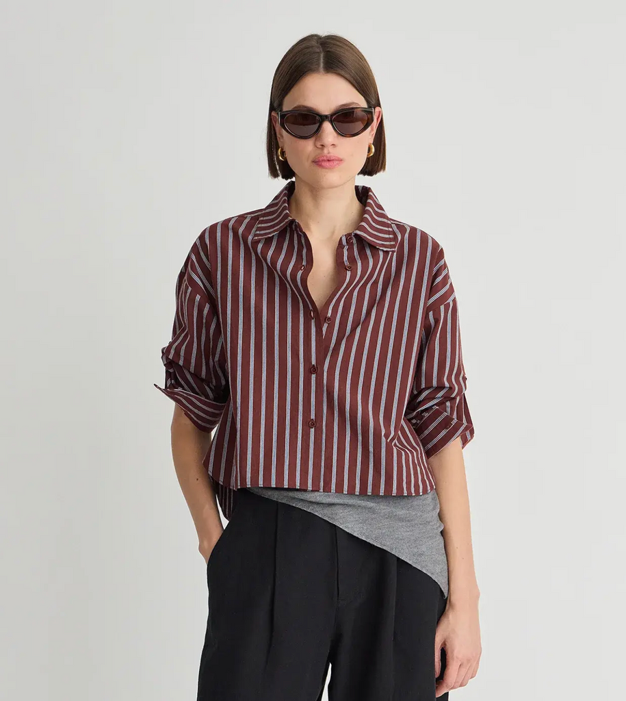 Woman wearing a burgundy and light blue vertically striped button-down shirt layered over a gray asymmetrical top, paired with black pleated trousers and dark sunglasses.