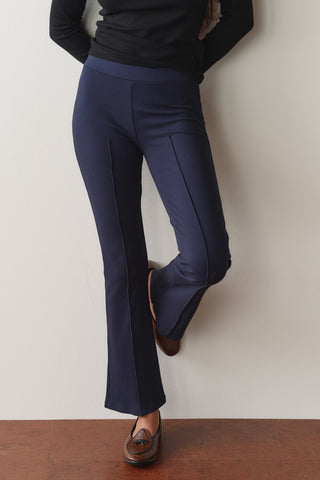Close-up of a person wearing DONNI navy blue ribbed knit kick flare pants with structured center pintuck seams, paired with a black long-sleeve top and brown leather tassel loafers against a neutral wall.