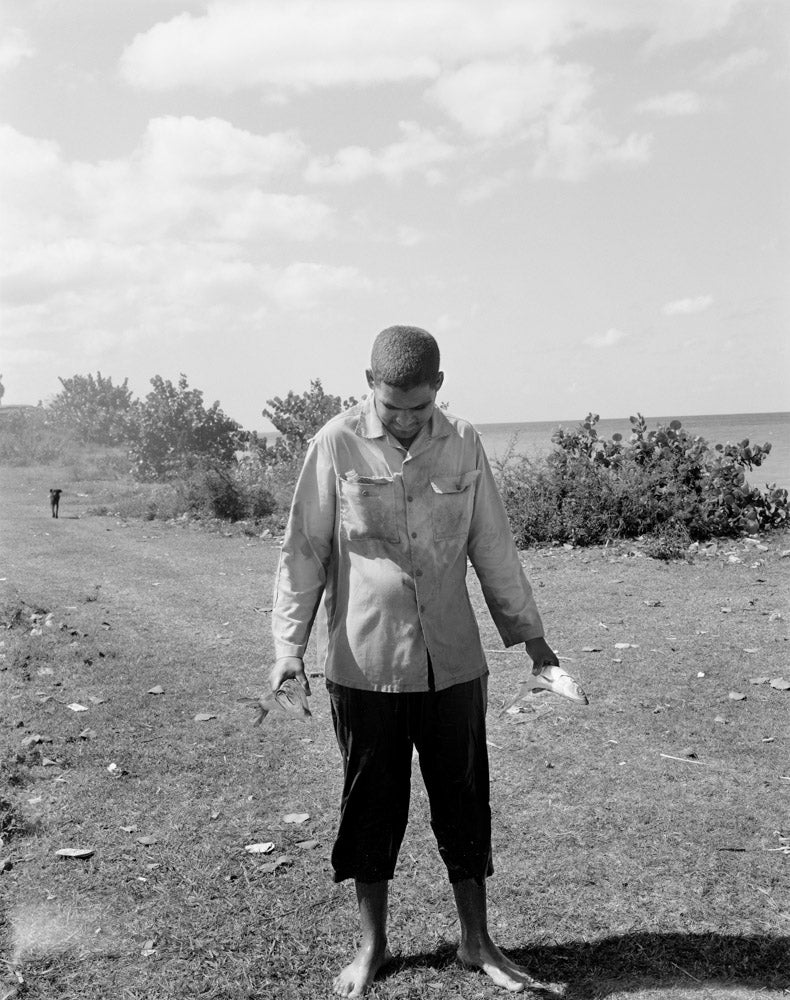 A black and white photograph of a young man barefoot on a grassy field near the ocean, looking down while holding a fresh fish in each hand. He is wearing dark cropped pants and a light-colored button-up shirt, with a body of water and a cloudy sky in the background.