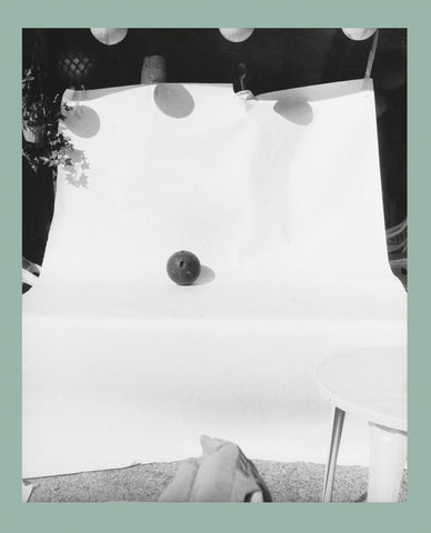 Black and white photograph featuring a dark, textured bowling ball centered on a large sheet of white paper backdrop.