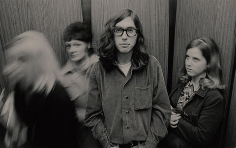 Black and white photograph of a man with long wavy hair and thick black glasses standing in the center of an elevator with wood paneling, surrounded by blurred figures of women.