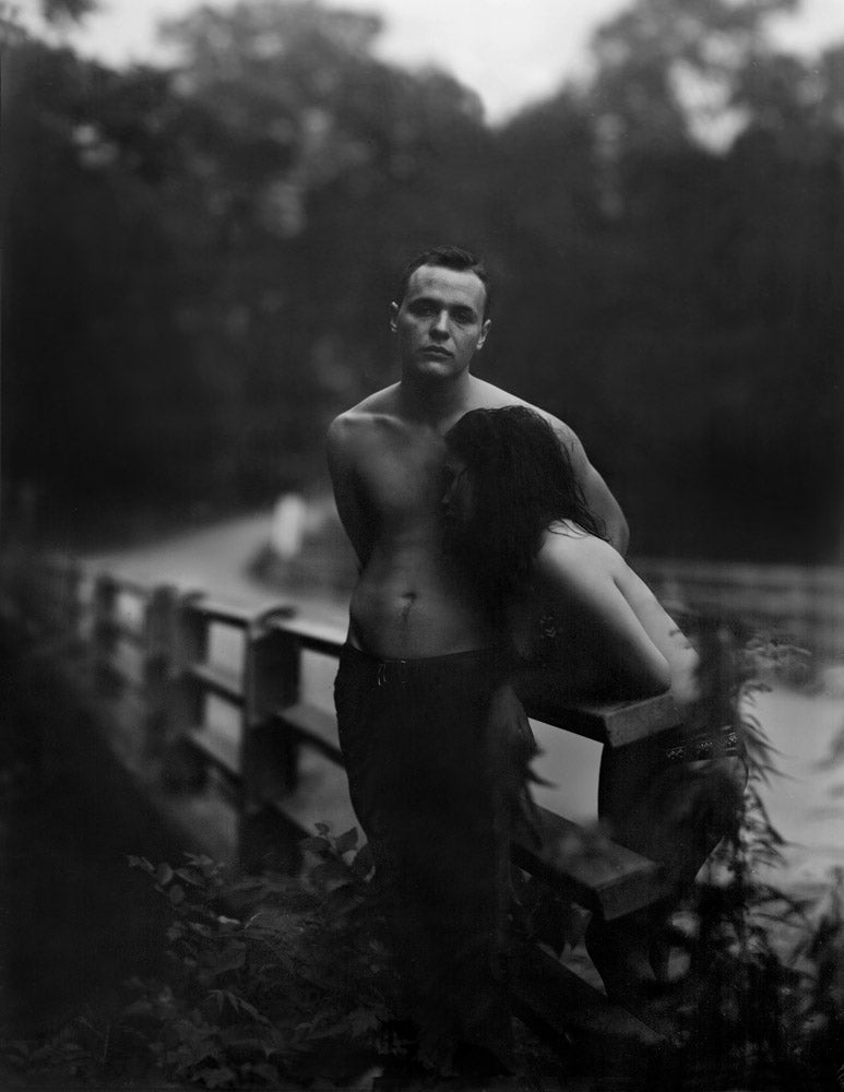 A moody black and white photograph by Raymond Meeks featuring a shirtless man standing on a rustic wooden bridge while a woman leans against him.